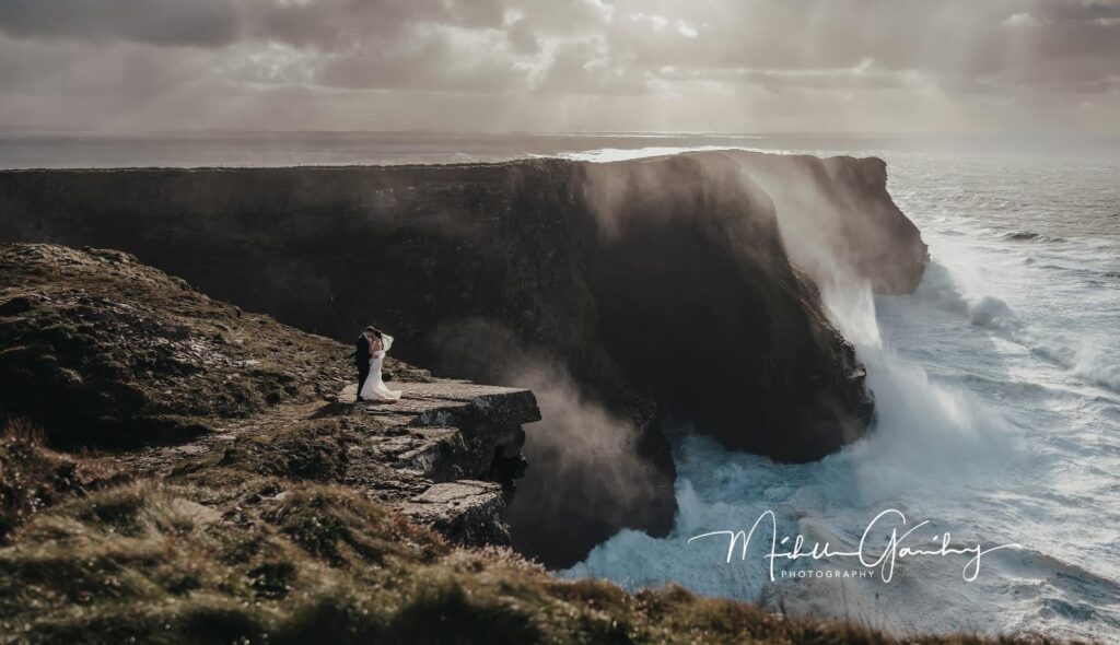 Couple embracing during their Cliffs of Moher elopement at Hags Head in Ireland with dramatic ocean waves crashing below the cliffs.