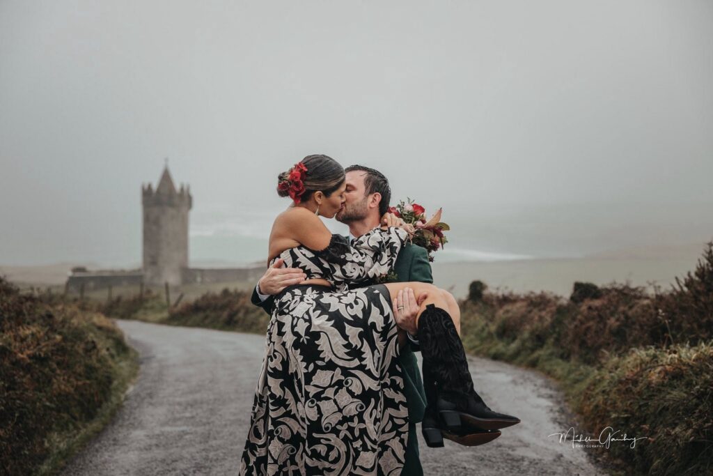 Couple sharing a romantic kiss on a country road in Ireland during their vow renewal, with a historic Irish castle in the background and the bride wearing a black and white floral dress.