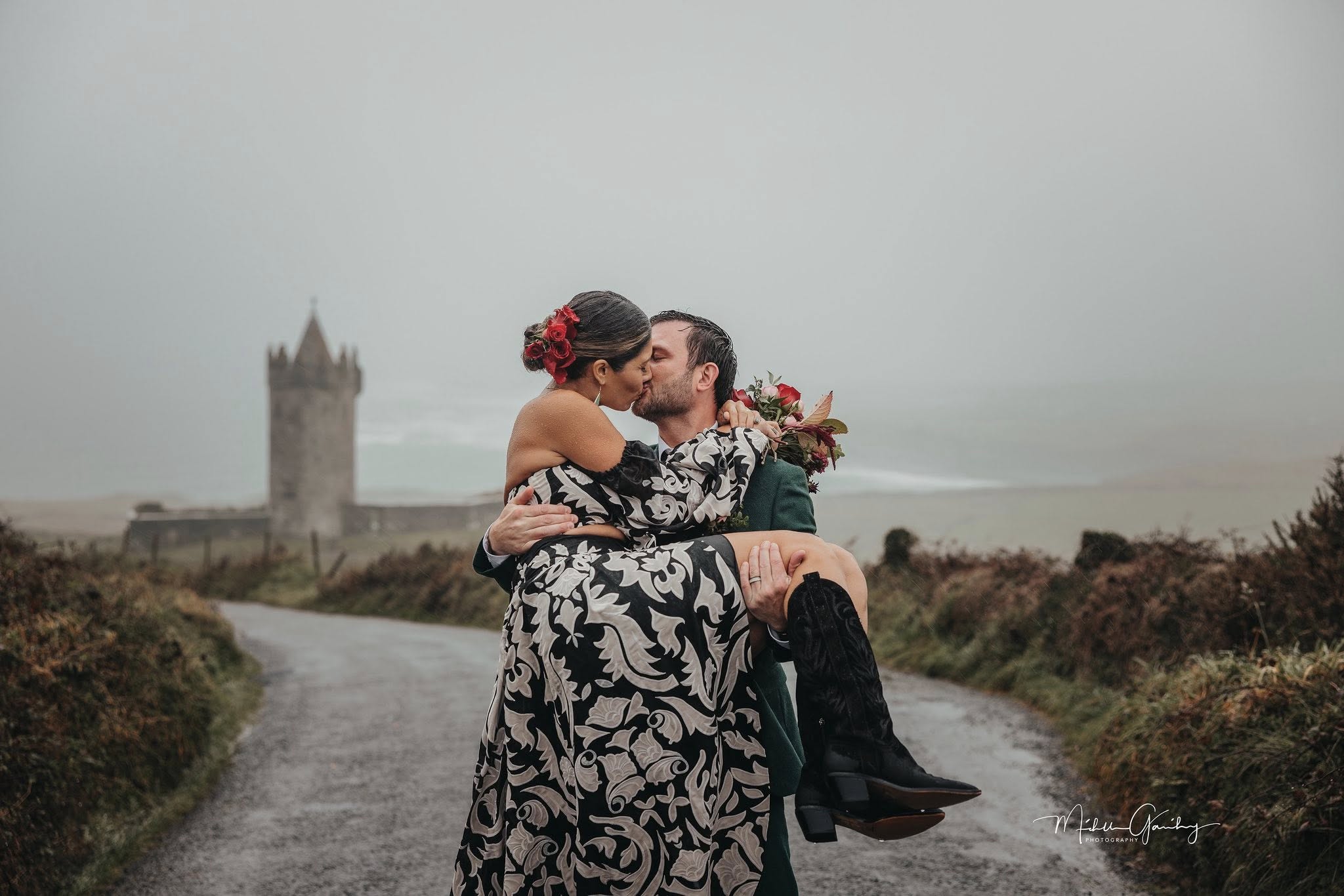 Couple sharing a romantic kiss on a country road in Ireland during their vow renewal, with a historic Irish castle in the background and the bride wearing a black and white floral dress.