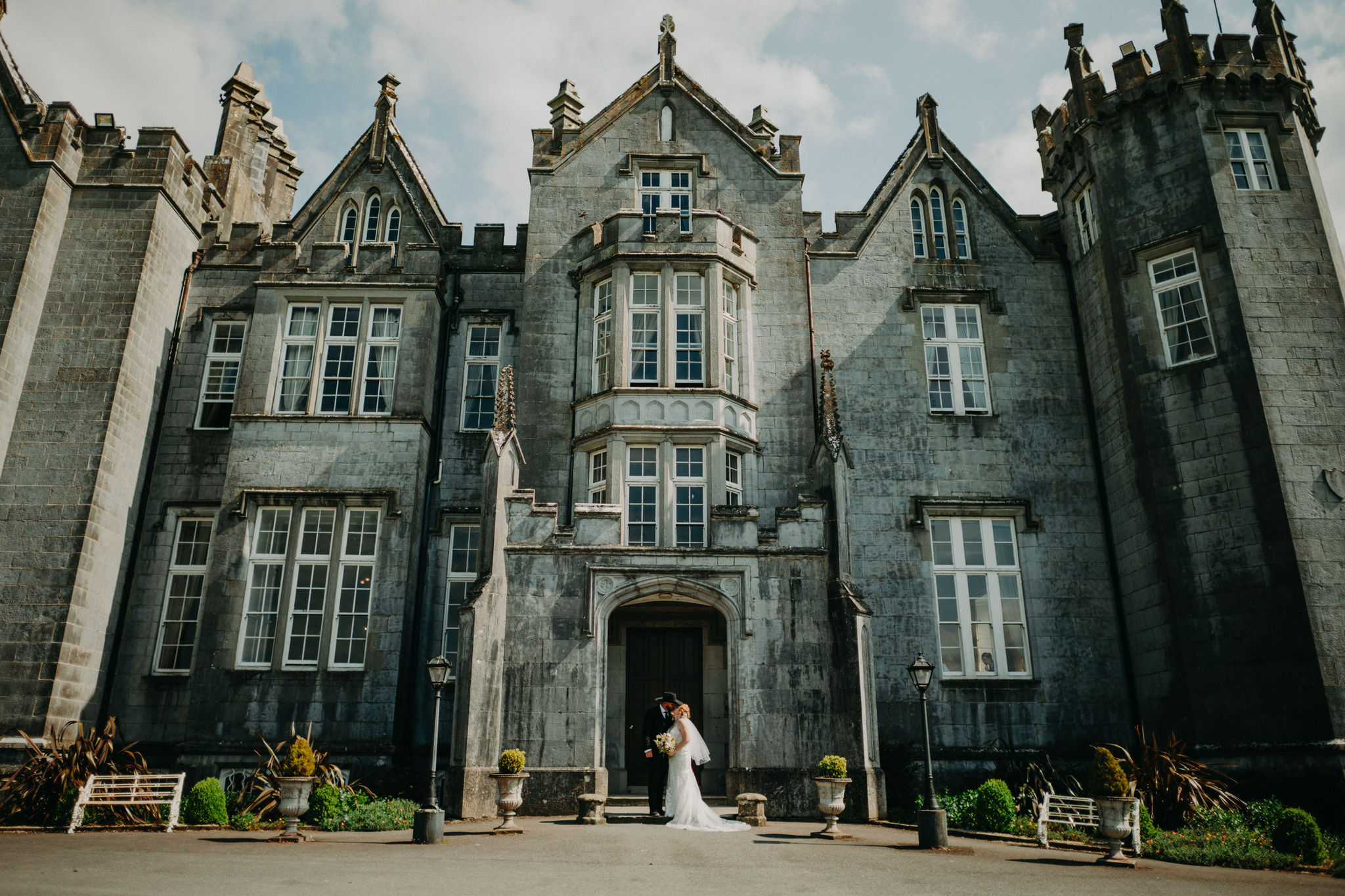 Newlywed couple standing outside the grand entrance of Kinnitty Castle in Ireland on a sunny day, with the historic stone castle towering above them.