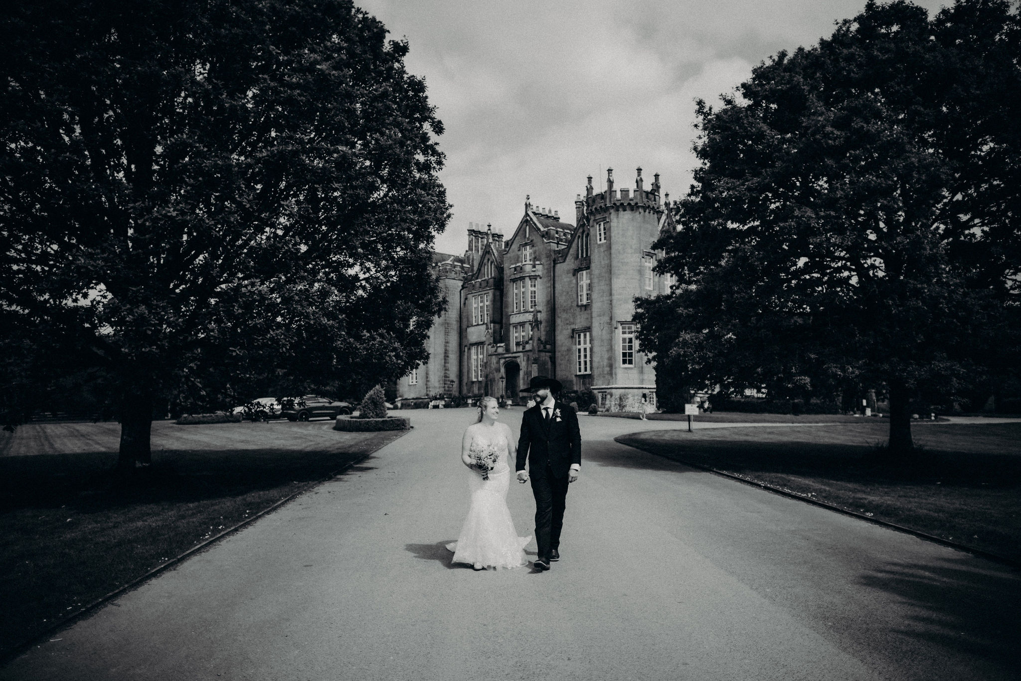 Black and white photo of a newlywed couple walking hand in hand along the driveway at Kinnitty Castle in Ireland.
