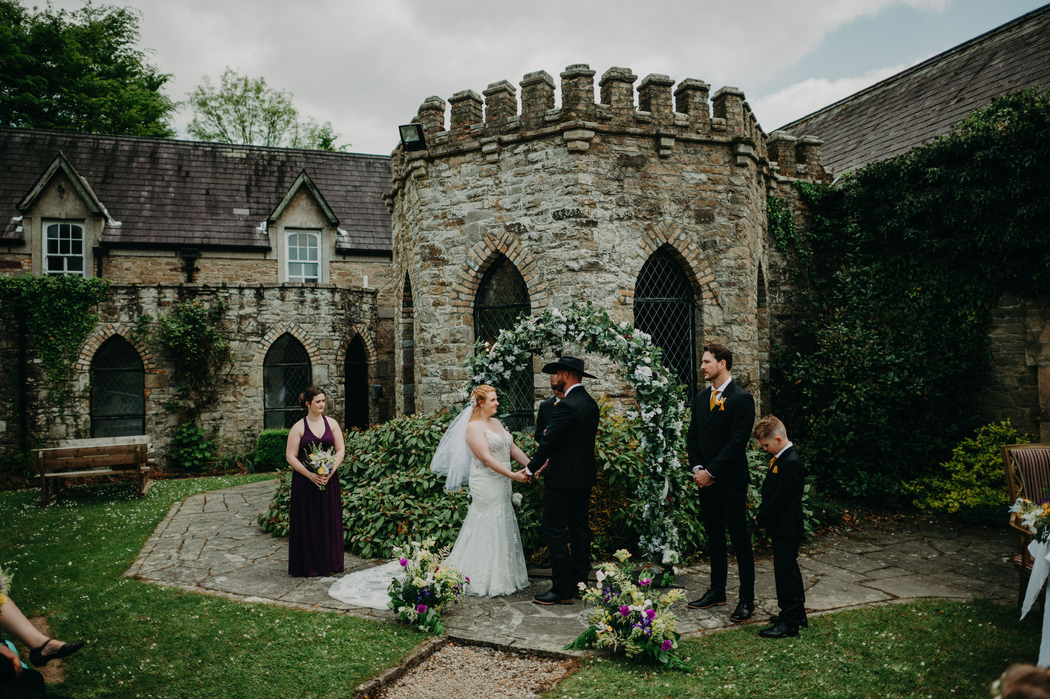 Outdoor wedding ceremony at Kinnitty Castle in Ireland with the couple standing beneath a floral arch in the castle courtyard.