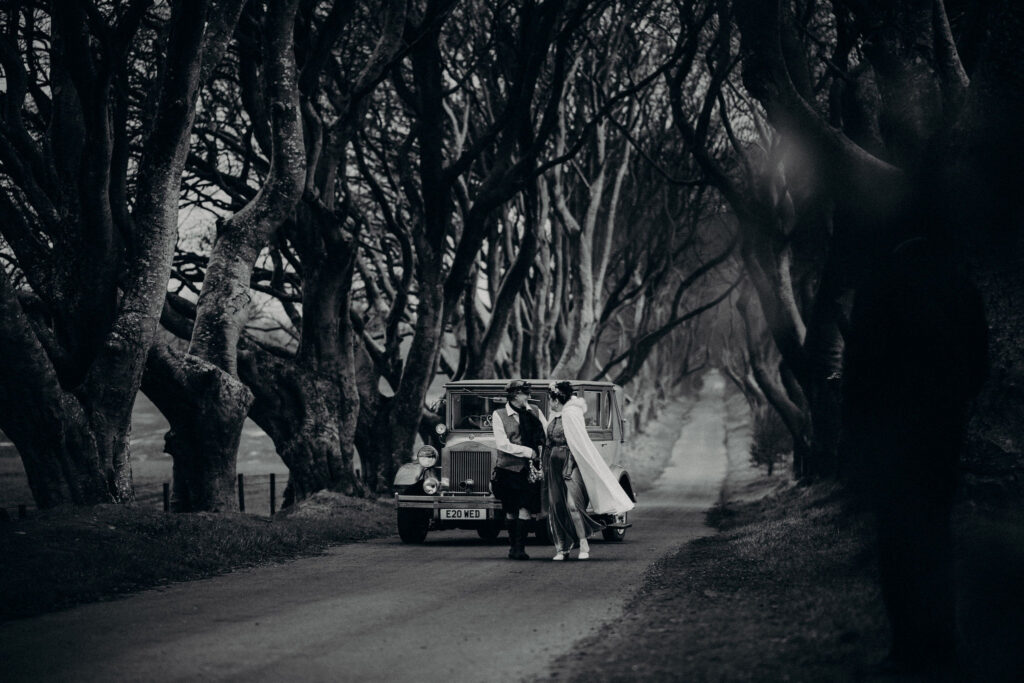 Couple standing beside a vintage car at The Dark Hedges in Northern Ireland during a destination wedding in Ireland, surrounded by dramatic twisting trees on a moody winter day.
