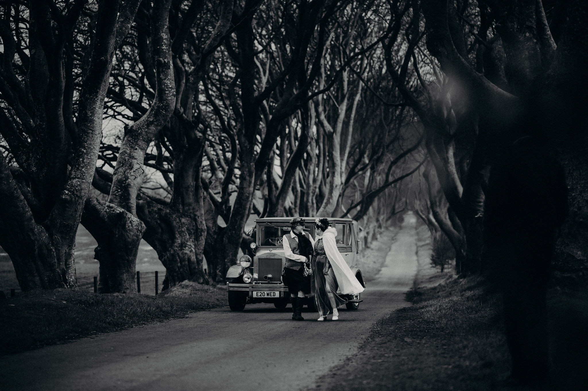 Couple standing beside a vintage car at The Dark Hedges in Northern Ireland during a destination wedding in Ireland, surrounded by dramatic twisting trees on a moody winter day.