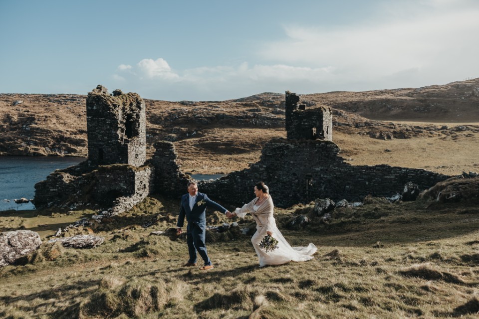 Adventure elopement couple running hand in hand beside ancient Irish ruins on a remote coastal landscape.