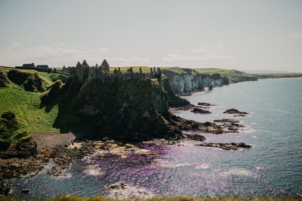 Dramatic coastal view of Dunluce Castle perched on rugged cliffs along Northern Ireland's Causeway Coast, overlooking the sparkling Atlantic Ocean and rocky shoreline below.