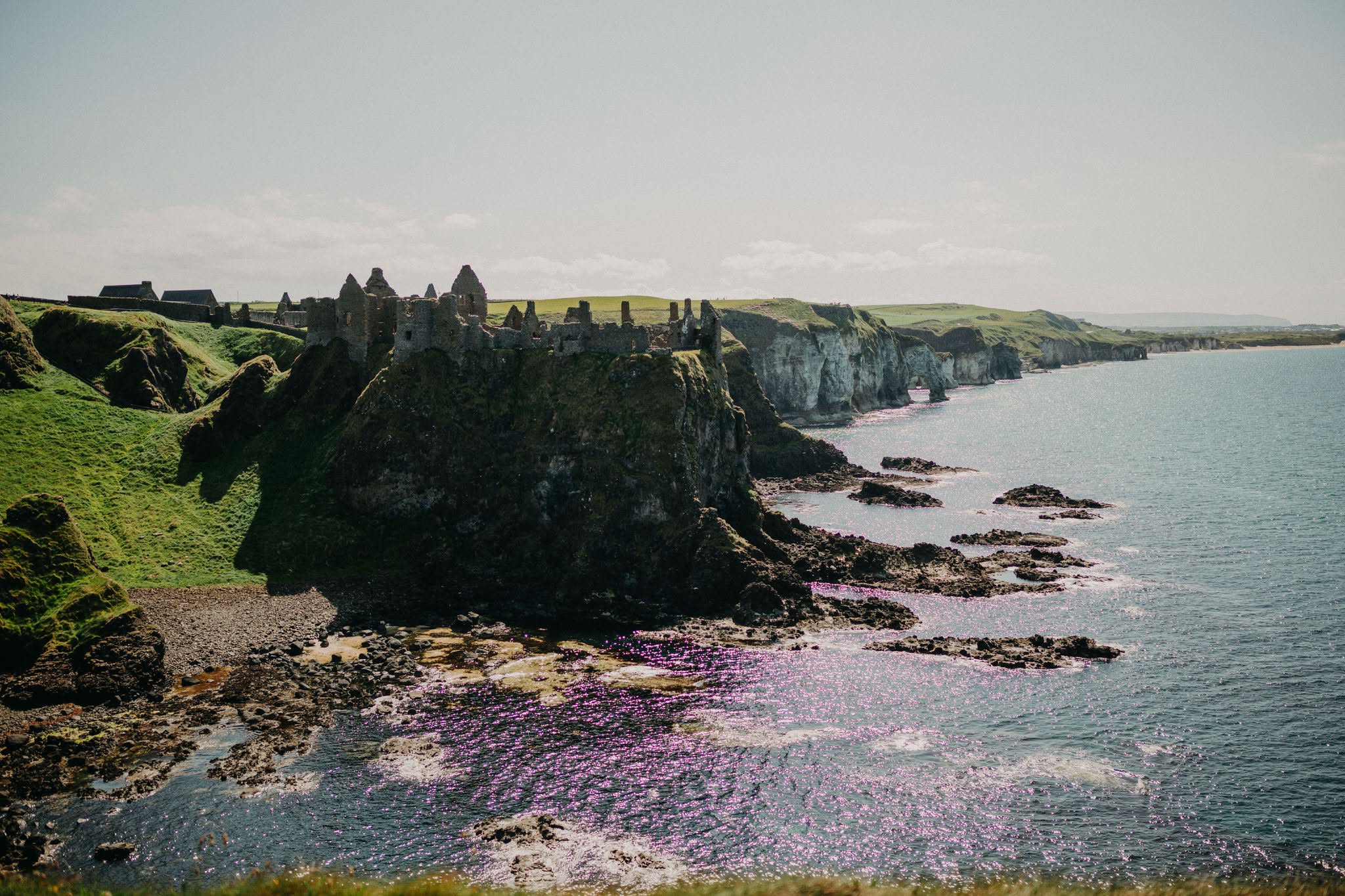 Dramatic coastal view of Dunluce Castle perched on rugged cliffs along Northern Ireland's Causeway Coast, overlooking the sparkling Atlantic Ocean and rocky shoreline below.