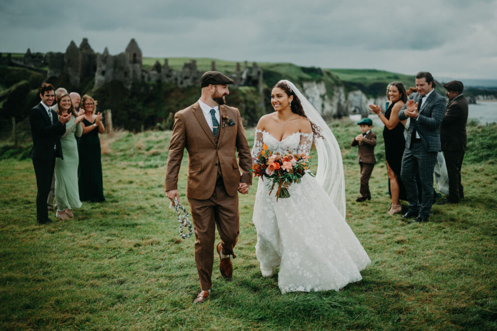 Newly married couple walking together as wedding guests applaud beside Dunluce Castle in Northern Ireland.