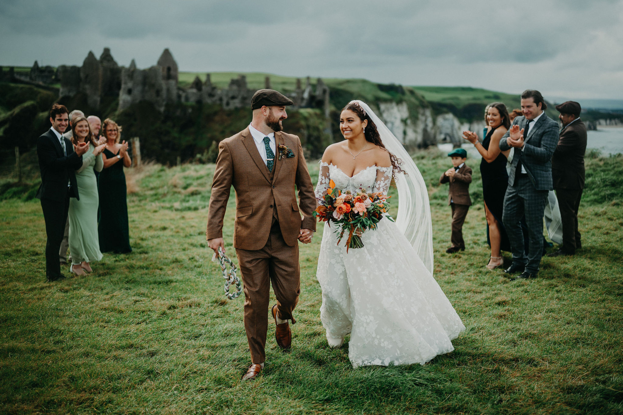 Newly married couple walking together as wedding guests applaud beside Dunluce Castle in Northern Ireland.